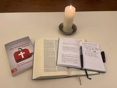 Buch "Der evangelische Patient" neben offener Bibel und Losung , Notizbuch, Stift und brennender Kerze